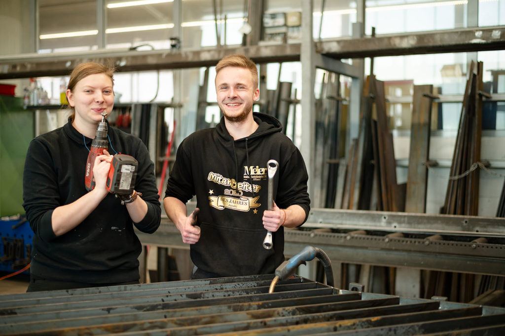 lächelnde Azubis Metallbau Meier mit Werkzeugen, Daumen hoch
